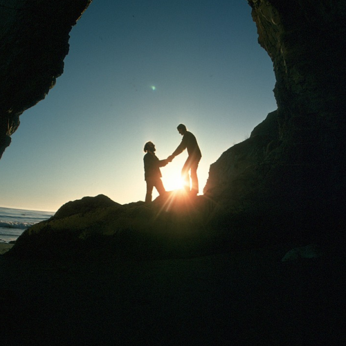 Zwei Menschen an einem Strand, man sieht nur die Silhouette. Ein Mensch gibt dem anderen Mensch unterstützend die Hand.